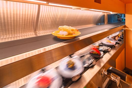 Long exposure photography of a bustling revolving sushi bar capturing the essence of the fast-paced Japanese dining with a plate of Deep fried shrimp tempura sushi delivered the above conveyor belt.