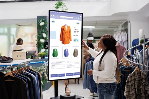 African american client selecting items to buy from self service checkout kiosk, reviewing modern clothes and sizes on interactive monitor. Young woman looking at products on board.
