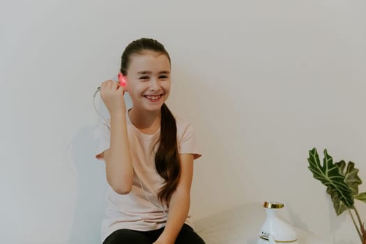 One beautiful Caucasian brunette girl with a happy smile, collected hair and in a pink T-shirt treats her ear with an infrared light device, sitting on her knees on a bed at home near a white wall, close-up side view.