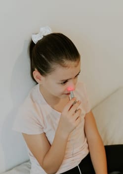 One beautiful Caucasian brunette girl with a serious emotion, collected hair and in a pink T-shirt treats the right nasal passage with a device with infrared light, sitting on the bed, cross-legged, leaning on a white wall, close-up view from above.