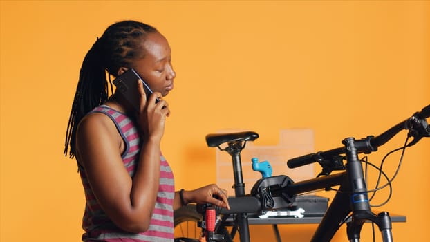 African american woman talking on phone with mechanic, asking for help on repairing damaged bike, studio background. BIPOC person on telephone call with bicycle engineer, camera A