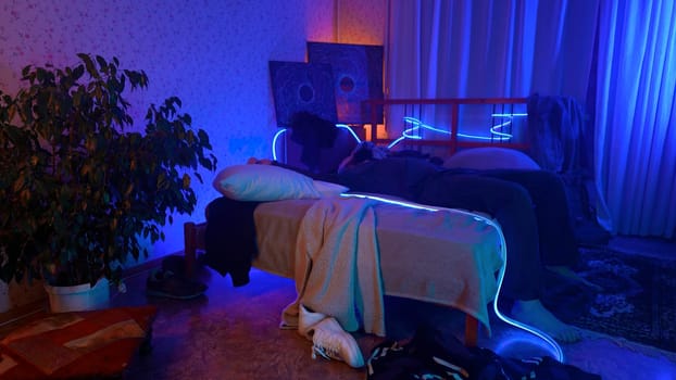 Worried, tired man sitting on bed at night and listening to music. Media. Young man can't sleep at night in a room with neon lighting