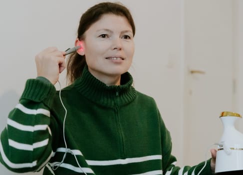 One beautiful Caucasian brunette girl with a smile on her face, gathered hair in a green sweater, treats her left ear with an infrared light device, sitting on a bed against a white wall, close-up view from below.