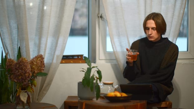 Young man drinks tea and looks at tablet in cafe. Media. Handsome young man is drinking tea alone and looking at tablet. Student is relaxing with tablet in cozy cafe.