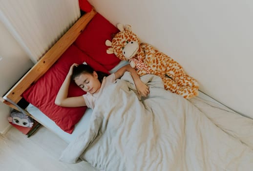 One beautiful little Caucasian brunette girl sleeps sweetly in a wooden bed on a red pillow with a soft toy giraffe, covered with a gray blanket, flat lay close-up.