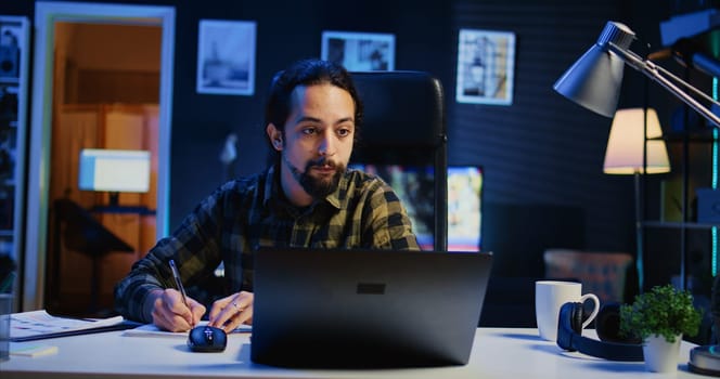 Remote employee working at home office desk, reading information on laptop and writing in notebook. Freelancer noting down ideas on paper using pen while teleworking, camera B