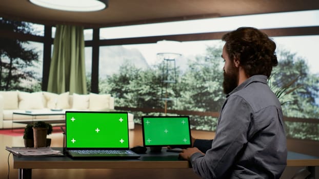 Businessperson using greenscreen displays on devices, sitting at his desk in luxurious villa in a forest. Rich entrepreneur looking at screens showing isolated copyspace, works on laptop and tablet.