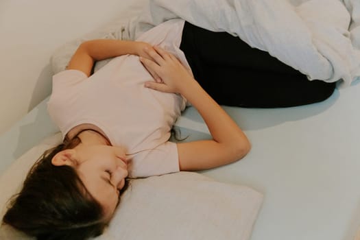 One beautiful little Caucasian brunette girl with flowing long hair sleeps in bed, curled up in pain and holding her stomach with both hands, close-up side view.