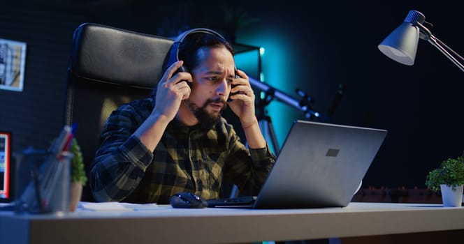 Freelancer enjoying music while researching data for company project, doing tasks in personal home office overnight. Remote worker sitting at desk in front of laptop and using headphones, camera B