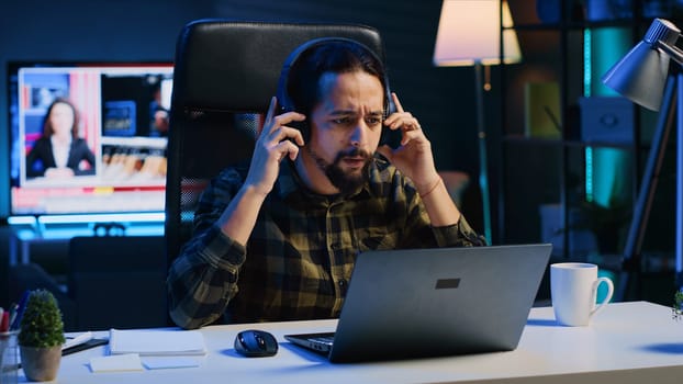 Freelancer enjoying music while researching data for company project, doing tasks in personal home office overnight. Remote worker sitting at desk in front of laptop and using headphones, camera A