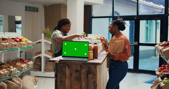 Vendor presenting eco goods on shelves next to greenscreen, recommends natural bulk products in organic shop. Laptop placed on stall running isolated chromakey layout template. Tripod shot.