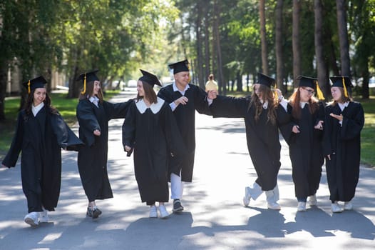 Row of happy young people in graduation gowns outdoors. Students are walking in the park