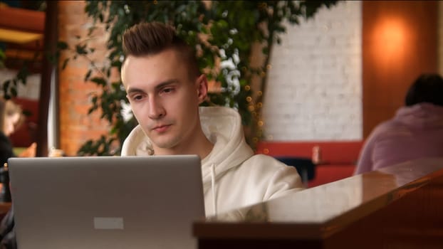 Young man is concentrating on laptop in cafe. Stock footage. Student is writing paper on laptop in cafe. Handsome young man freelancing at laptop in cafe.