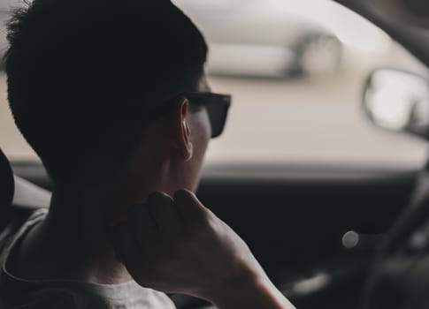 Portrait of a young handsome caucasian brunette guy with a beard and sunglasses thoughtfully sitting sideways behind the wheel of a car in a traffic jam, holding his hand near his neck and looking in the rearview mirror, bottom view close-up.