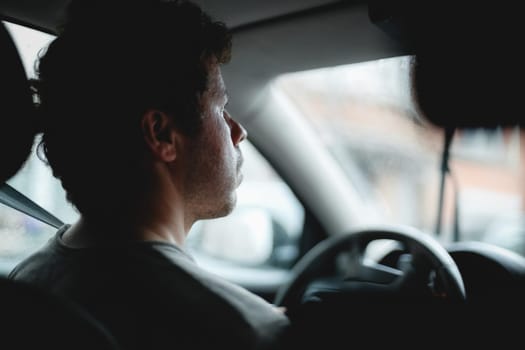 Young handsome caucasian guy with brown curly hair carefully looks behind the road while driving a car on a rainy day, traveling around the city, side view close-up in dark style and selective focus. The concept of driving a car, taxi services, travel, city life, solo trip, modern lifestyle.