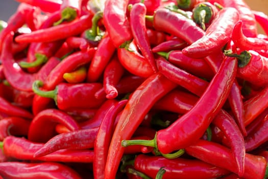 A vibrant assortment of fresh red peppers is seen heaped together, displayed at an outdoor market in broad daylight, showcasing their bright red hue and green stems.