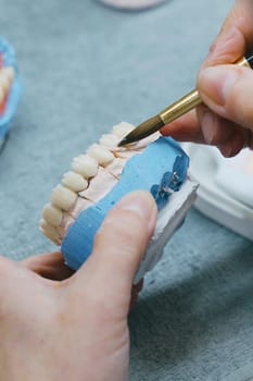 A close-up of a dental technician holding a toothbrush in their right hand, part of the process of manufacturing dental prosthesis. Vertical photo.
