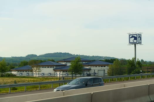 Weimar, Germany - May 7, 2023: A car travels on a highway by modern buildings with blue roofs, as green hills stretch out under a clear sky in the background.