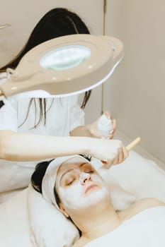 A young beautiful Caucasian brunette girl, a cosmetologist, smears a white cream mask with a brush on the face of an adult female client in the cheekbone area, who is lying with her eyes closed on a massage table in a beauty salon, close-up top view.