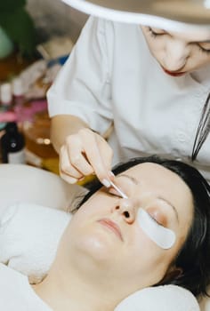 Portrait of a young beautiful Caucasian brunette cosmetologist girl in a white coat who degreases eyelashes on the lower eyelid with a cotton swab to a woman lying on a cosmetology table in a home beauty salon, close-up side view.