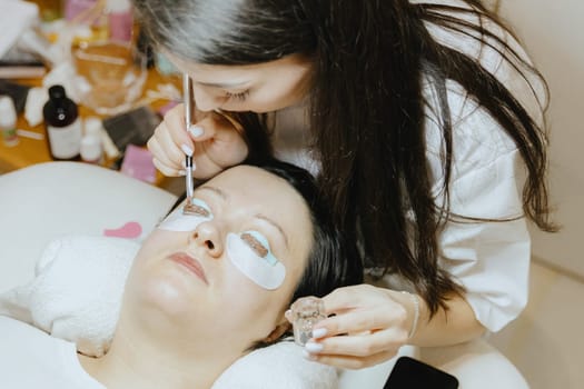 Portrait of a young semi-recognizable Caucasian girl cosmetologist in a white coat applying brown eyelash paint on a silicone mold of the right eye using a brush to a brunette woman lying on a cosmetology table in a home beauty salon for eyelash lift, top side close-up view with selective focus.