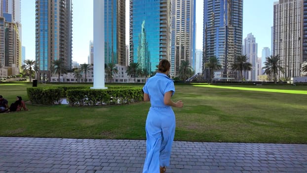 Rear view of a young woman running on stone path in a city park, Dubai. Action. Woman spending time outdoors near tall buildings