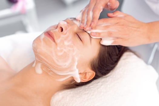 Cosmetologist cleanses the skin with foam. Woman's skin care procedure in beauty clinic. Natural cosmetics. Cosmetic treatment of face. Therapeutic peeling cream.