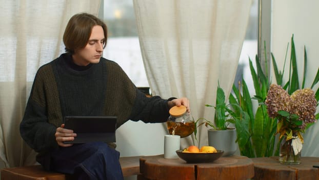 Young man is sitting in cafe with tea and tablet. Media. Man in cozy cafe is working on tablet and drinking tea. Young freelancer works in cafe with tablet.