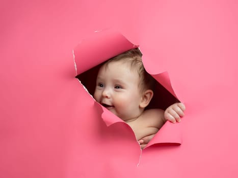 Cute Caucasian newborn baby boy peeks out of a hole in a paper pink background