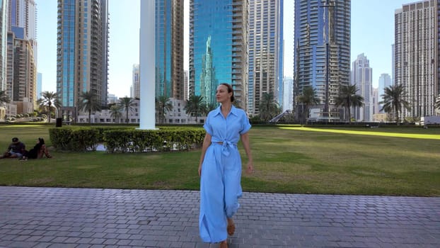 Happy woman in blue summer suit walking in city park. Action. Green meadow and skyscrapers on the background