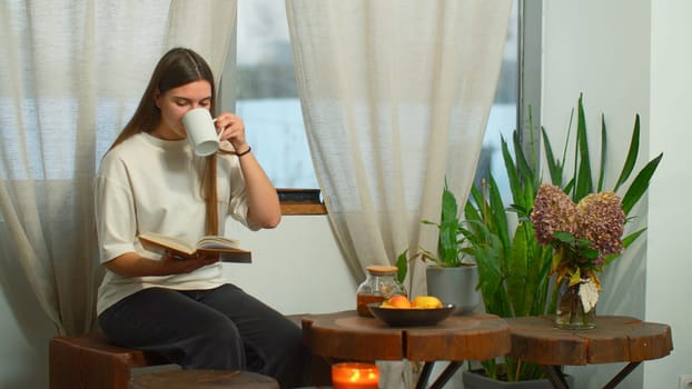 Young woman is reading book with tea in cozy cafe. Media. Beautiful young woman is reading book in cafe. Cozy winter day in cafe with mug of tea and favorite book.