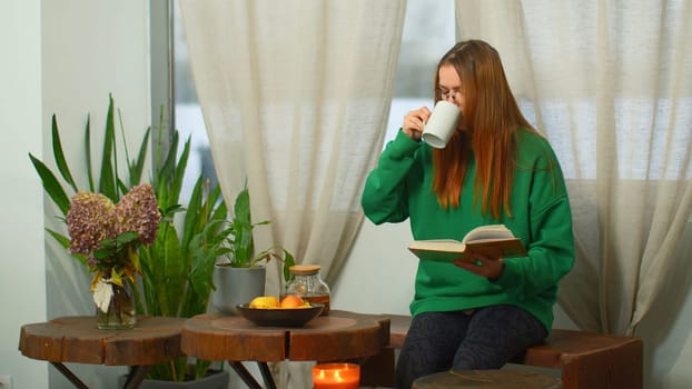 Young woman with glasses is reading book in cafe. Media. Beautiful female student is reading book in college cafe. Smart student studies and reads books in cozy cafe.