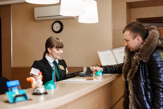Side view of female receptionist and male guest standing at hotel reception and paying with check during check-in