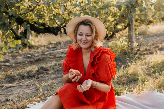 A woman in a red dress is sitting in a field and eating a piece of fruit. She is wearing a straw hat and she is enjoying the outdoors