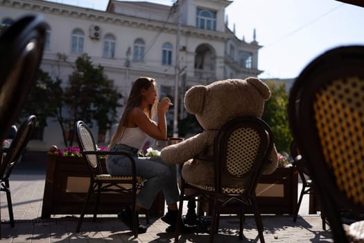 A woman sits cafe with a teddy bear next to her. The scene is set in a city with several chairs and tables around her. The woman is enjoying her time at the outdoor cafe