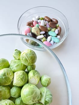 Brussels sprouts in clear glass bowl above assorted candy in another bowl on white surface. Healthy eating concept. High quality photo