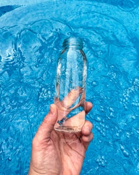 Hand holding clear glass bottle partially filled with water against sparkling blue water background. Clean water concept. High quality photo