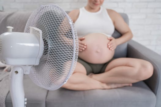 A pregnant woman is sitting on the couch, stroking her tummy and enjoying the cool air from an electric fan