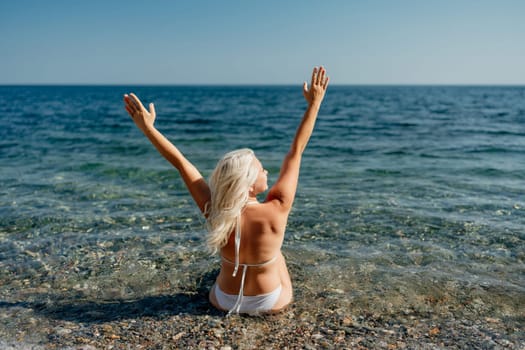 A woman is sitting in the ocean, with her arms raised in the air. She is wearing a bikini and she is enjoying the water