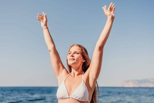 A woman in a bikini is standing on the beach and raising her arms in the air. Concept of freedom and joy, as the woman is celebrating or expressing happiness