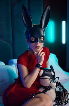 sexy girl in a bdsm rabbit mask and a bright red dress is sitting on a bed with a teddy bear in shoulder straps and leather belts in a neon light