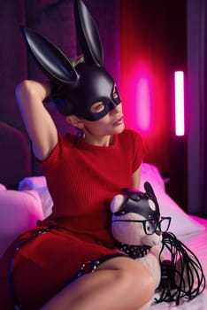 sexy girl in a bdsm rabbit mask and a bright red dress is sitting on a bed with a teddy bear in shoulder straps and leather belts in a neon light