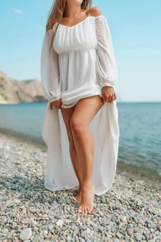 A woman in a white dress is standing on a beach. The dress is long and flowing, and the woman is holding it up to her knees. The beach is rocky and the water is calm. The scene is serene and peaceful