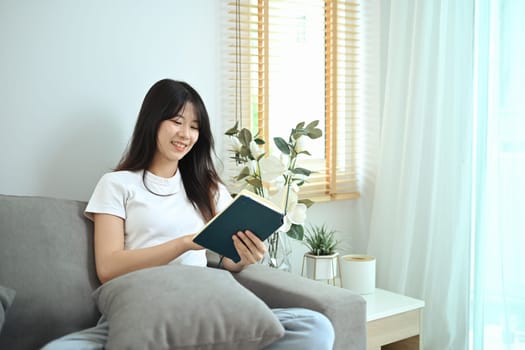 Happy young woman wearing casual clothes relaxing in her living room and reading book.