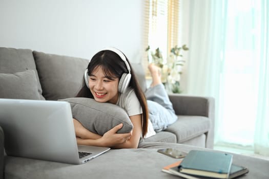 Cheerful asian woman in wireless headphones using laptop while lying on couch.