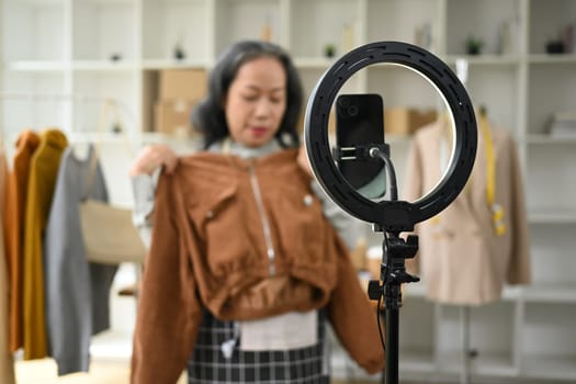 Smiling middle age woman showing and review product in front of smartphone live streaming at her shop.