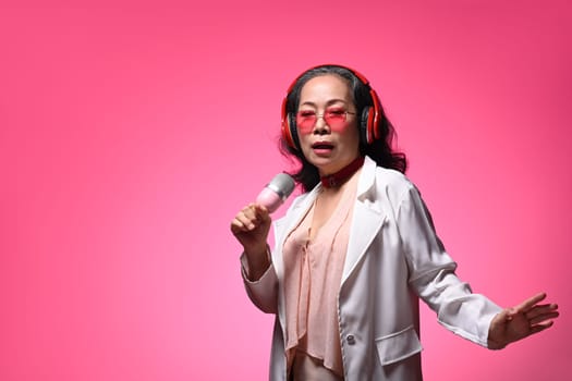 Stylish middle age lady in headphone singing with microphone on pink background.