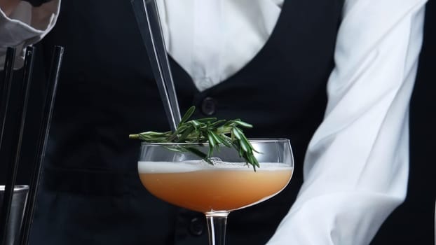 Close up of smart bartender prepare cocktail with orange and rosemary at bar and modern pub. Professional bartender hands making a blood orange negroni with black background. Slow motion. Comestible.