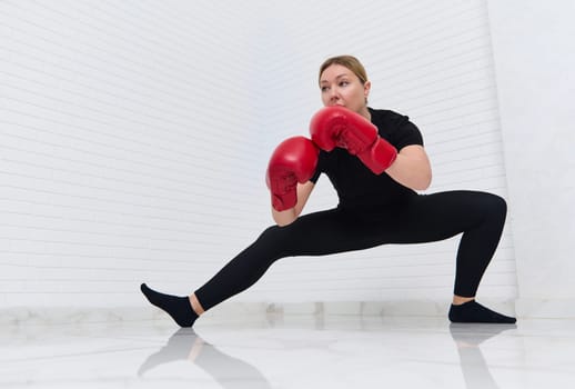 European woman fighter boxer in red boxing gloves, doing stretching exercises on legs,warming up, getting for combat, match, challenge, isolated over white background. Martial art. People and sport.
