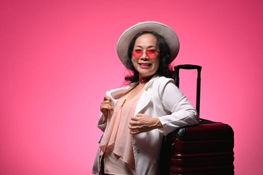 Cheerful retired woman tourist standing with suitcase isolated on pink background. Summer, travel and vacation concept.